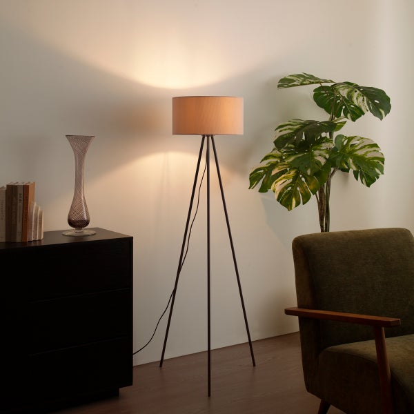 Dreibein-Stehleuchte mit Textilschirm in einem wohnlich eingerichteten Zimmer neben einer Pflanze und einem Sessel.