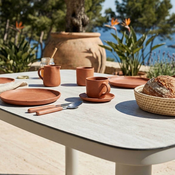 Hell gedeckter Gartentisch im mediterranen Stil mit Terrakotta-Geschirr, Besteck und Brotkorb im sonnigen Außenbereich.