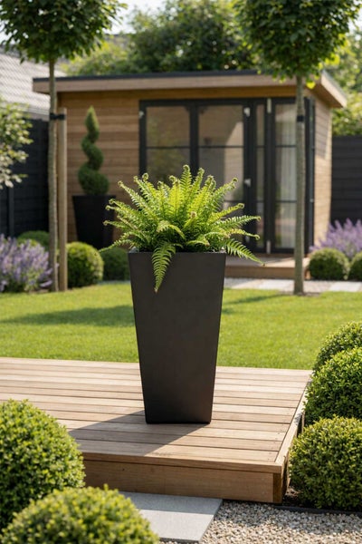 Hoher, quadratischer Pflanzkübel mit Farn auf einer Holzterrasse in einem hellen, modern gestalteten Garten mit Gartenhaus.