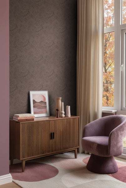Wohnzimmer mit dunkelbrauner Blumentapete, Sideboard aus Holz, lila Sessel und gemustertem Teppich vor einem Fenster.