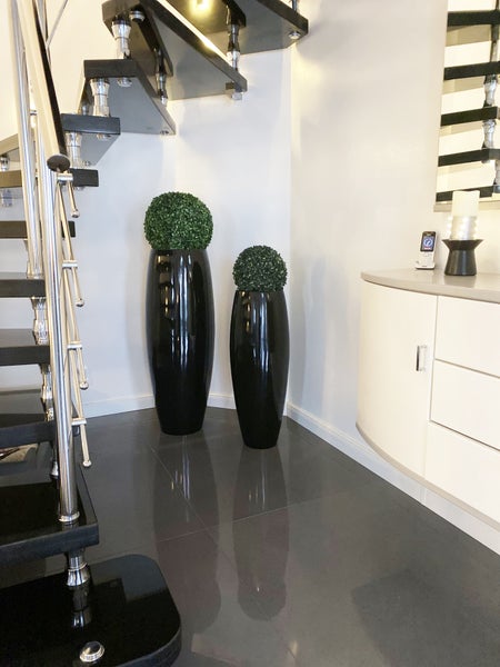 Moderner Flur mit schwarzer Treppe, hohen glänzenden Bodenvasen mit Buchsbaumkugeln und einem hellen Sideboard auf dunklem Fliesenboden.