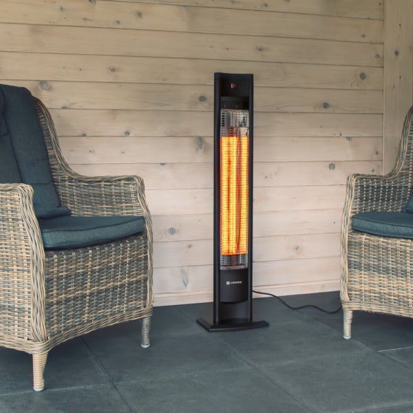 Schwarzer Infrarot-Terrassenstrahler zwischen zwei Rattansesseln auf einer Terrasse mit Holzwand und Fliesenboden.