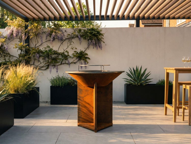 Helle Terrasse mit Feuerschale und Grill aus Cortenstahl. Hintergrund mit Holzpergola, Glyzinien an weißer Wand, schwarzen Pflanzgefäßen und Steinfliesen.