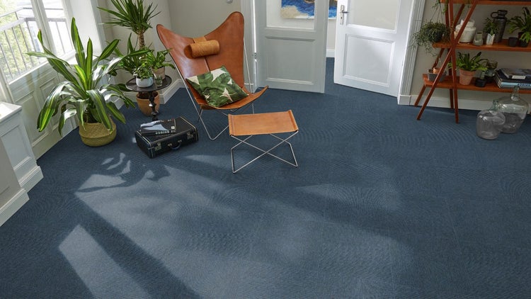 Helles Wohnzimmer mit blauen Teppichfliesen, Leder-Schmetterlingsstuhl mit Hocker, Zimmerpflanzen und Holzregal.