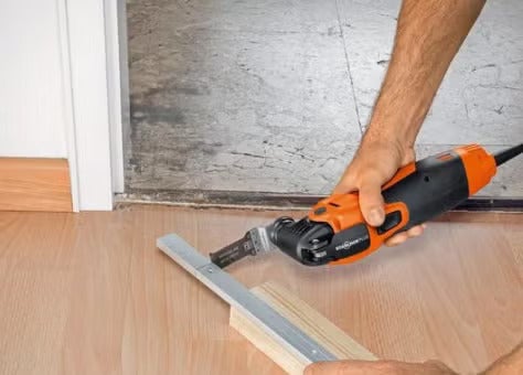 Eine Hand hält ein Multifunktionswerkzeug beim Kürzen eines Türzargenabschlusses auf einem Holzfußboden.