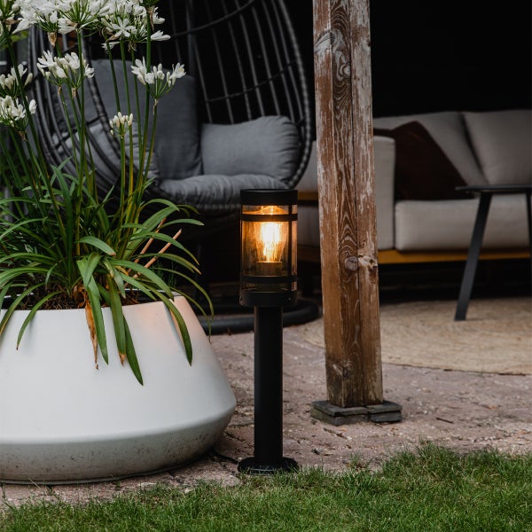 Schwarze Außen-Sockelleuchte mit warm leuchtendem Filament-Leuchtmittel auf einer Terrasse neben einem großen weißen Pflanzkübel und Gartenmöbeln.