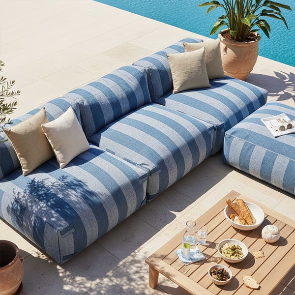 Modulare Gartenlounge mit blau-weiß gestreiftem Bezug, passendem Hocker und Holztisch in einer hellen Terrassenszene am Pool.
