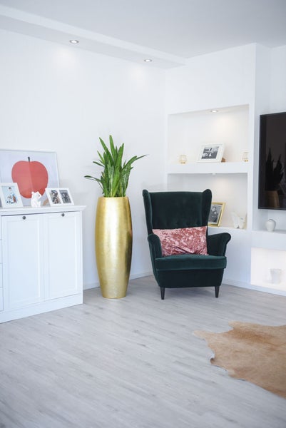 Modernes helles Wohnzimmer mit dunkelgrünem Samtsessel, hoher goldener Bodenvase mit Pflanze und weißem Sideboard auf hellem Bodenbelag.