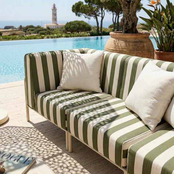 Gestreiftes Outdoor-Sofa mit Kissen auf einer hellen Terrasse neben einem Pool, mit einem Leuchtturm im Hintergrund.