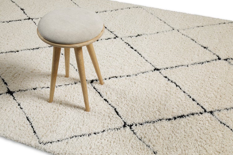 Hochflor-Teppich mit schwarzem Rautenmuster und ein hölzerner Hocker mit gepolsterter Sitzfläche.