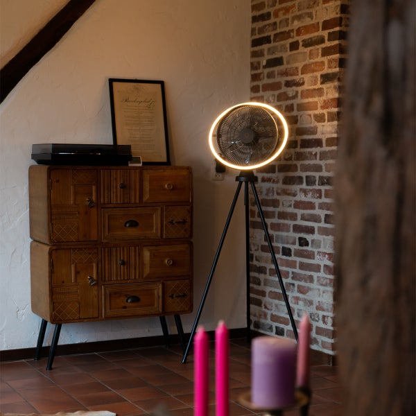 Moderner Stativventilator mit integriertem LED-Lichtring in einem rustikalen Wohnzimmer neben einer Kommode aus Holz und einer Backsteinwand.