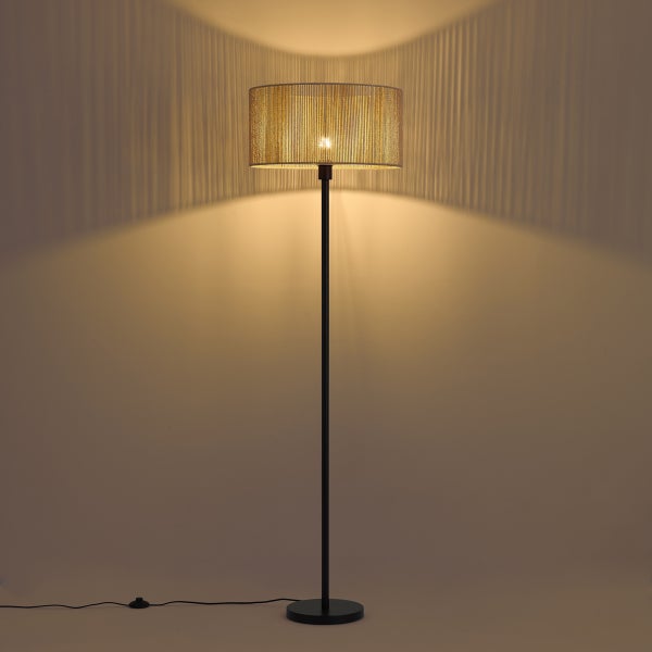 Stehlampe mit Metallgestell und rundem Schirm aus feinen Fäden, eingeschaltet mit Lichteffekt an der Wand.