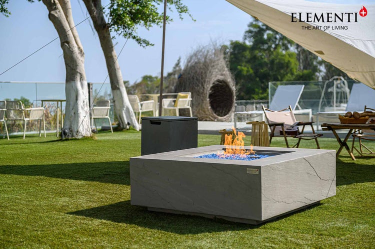 Quadratische Feuerstelle in Steinoptik mit blauen Glassteinen und Flammen in einem hellen Garten mit Sitzbereich, Elementi Logo