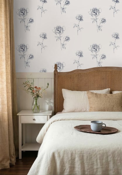 Schlafzimmer mit Vliestapete mit Rosenmuster über einer weißen Wandpaneele, Bett mit Rattankopfteil und weißem Nachtschrank.