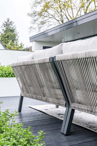 Modernes Gartensofa mit Seilgeflecht-Rückenlehne, dunklem Metallgestell und hellen Polstern auf einer Terrasse.