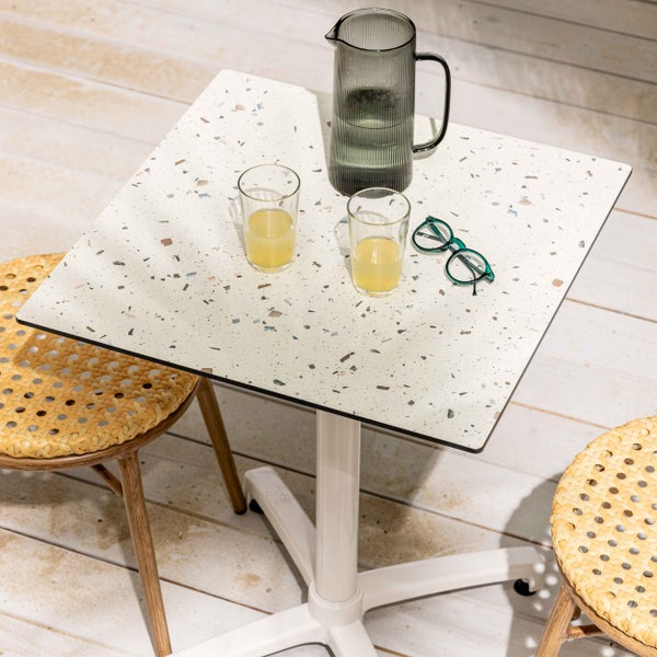 Quadratischer Bistrotisch mit Tischplatte in Terrazzo-Optik und weißem Mittelfuß, umgeben von geflochtenen Stühlen auf einer hellen Holzterrasse.