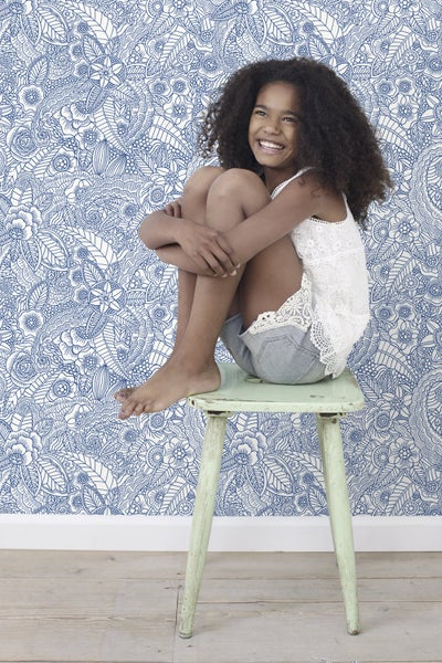 Mädchen sitzt vor einer Wand mit gemusterten Tapeten auf einem Hocker