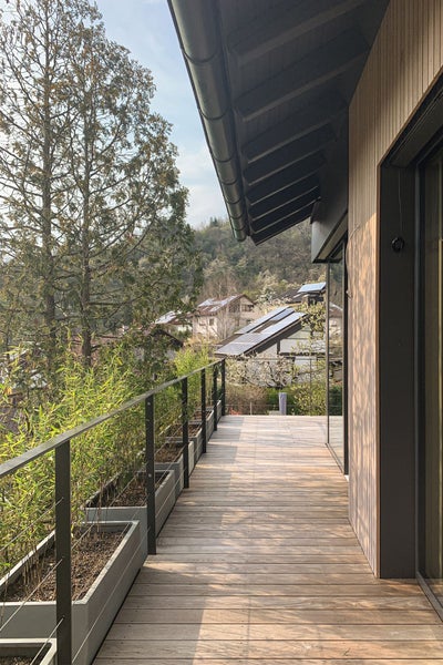 Balkon mit Holzterrasse, Metallgeländer, grauen Pflanzkübeln mit Bambus und Blick auf eine Hügellandschaft.