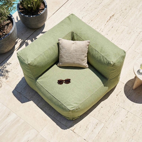 Grünes Eck-Loungekissen aus Stoff für den Außenbereich mit beigem Zierkissen und Sonnenbrille auf einer hellen Steinterrasse.