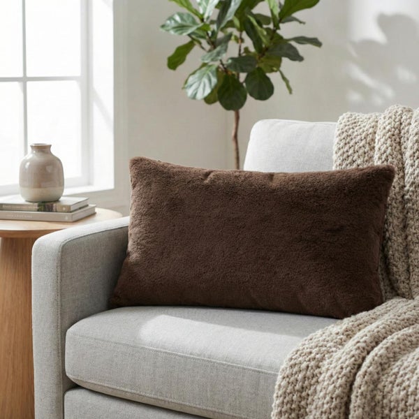 Rechteckiges braunes Plüschkissen auf einem hellen Sessel neben einer groben Strickdecke in einem hellen Wohnzimmer.