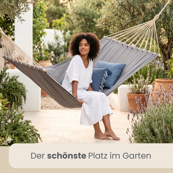 Frau sitzt entspannt in einer Hängematte mit Kissen in einem hellen Garten mit Terrakottatöpfen und Lavendel.
