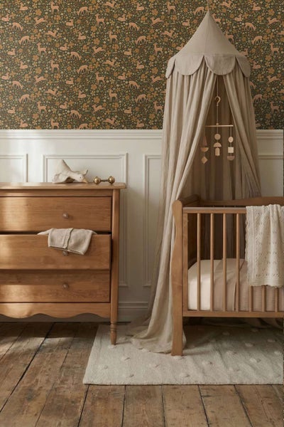 Ein gemütliches Babyzimmer mit Holzgitterbett, Betthimmel und Kommode vor einer Tapete mit Tiermotiven und Blumen auf einem Holzboden.
