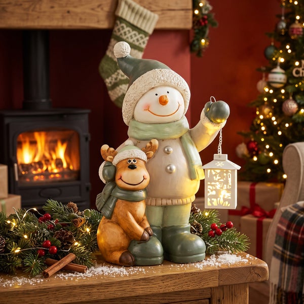 Dekorative Schneemannfigur mit Rentier und beleuchteter Laterne als Weihnachtsdekoration auf einem Holztisch vor einem Kamin.