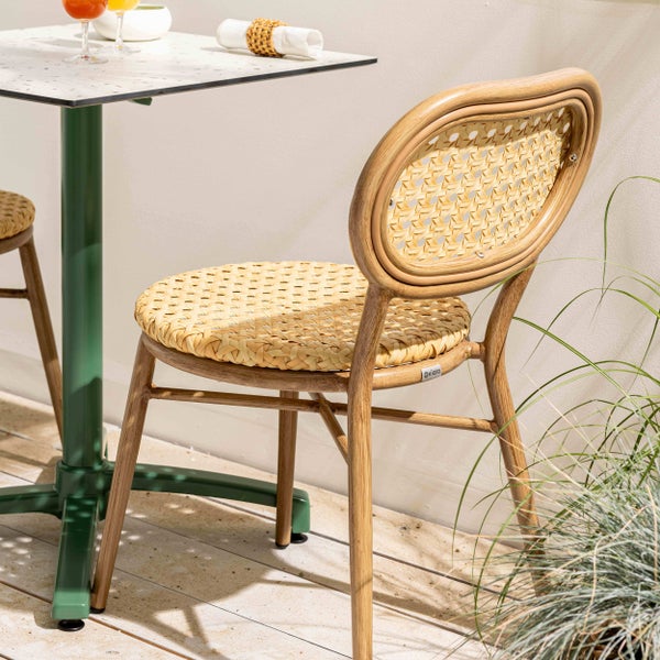 Moderner Bistrostuhl mit Geflechtsitz und Rückenlehne in Rattan-Optik auf einem Gestell in Holzoptik neben einem grünen Gartentisch.