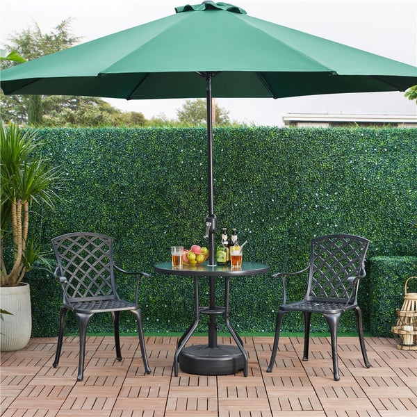Grüner Sonnenschirm über einem runden schwarzen Metalltisch und zwei passenden Stühlen mit Gittermuster auf einer Holzterrasse vor einer Hecke.