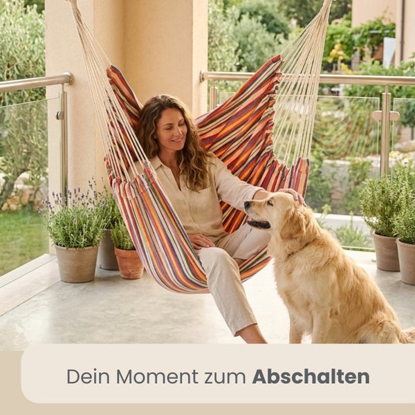 Frau sitzt in gestreiftem Hängesessel auf hellem Balkon mit Golden Retriever und Topfpflanzen. Moderne Atmosphäre mit Glasgeländer.