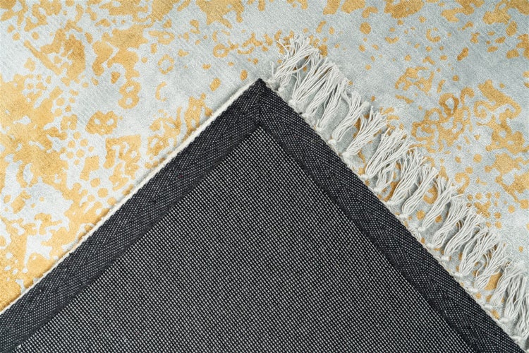 Teppich-Detail mit abstraktem Muster in Gold und Grau, hellen Fransen und dunkler Rückseite.