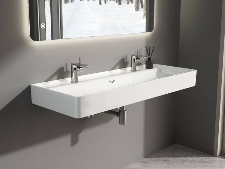 Modernes Doppelwaschbecken aus weißer Keramik zur Wandmontage mit zwei verchromten Armaturen unter einem beleuchteten Spiegel.