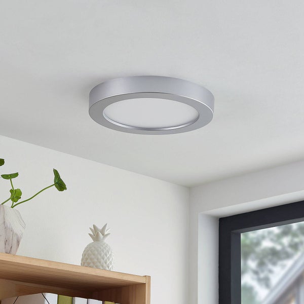 Flache, runde LED-Deckenleuchte mit silbernem Rahmen an einer Decke über einem Holzregal in einem hellen, modern eingerichteten Zimmer.