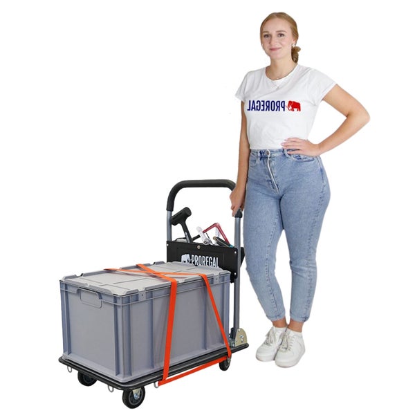 Eine junge Frau steht neben einem Rollwagen mit einer grauen Transportbox, die mit einem orangefarbenen Gurt befestigt ist. Werkzeuge befinden sich in einer Halterung am Rollwagen.