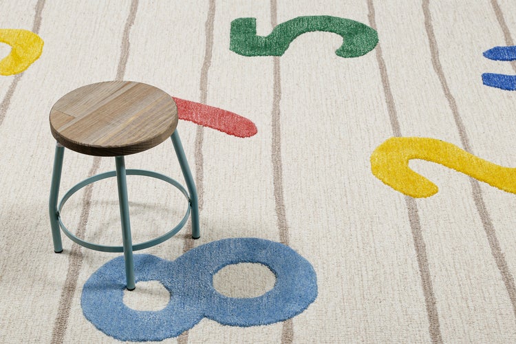Bunter Kinderteppich mit Zahlenmotiven und Linien, darauf ein Holzhocker mit Metallgestell.