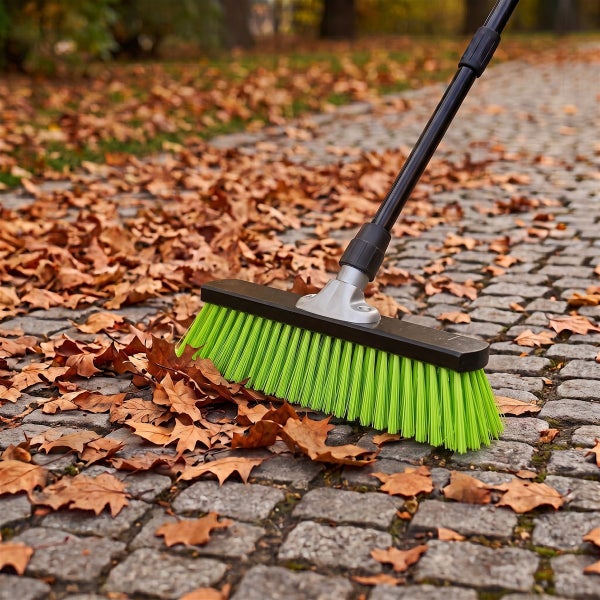 Ein Straßenbesen mit Stiel kehrt herbstliches Laub von einem gepflasterten Gehweg.