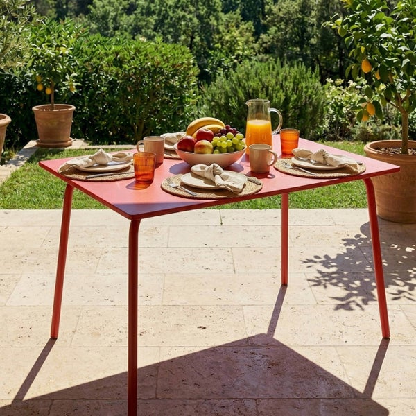 Gedeckter roter Gartentisch auf einer sonnigen Terrasse mit Steinboden vor einem grünen Garten mit Zitronenbäumen.