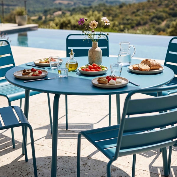 Runder Gartentisch mit vier passenden Metallstühlen auf einer hellen Terrasse am Pool, gedeckt mit Tellern, Gläsern und einer Blumenvase.