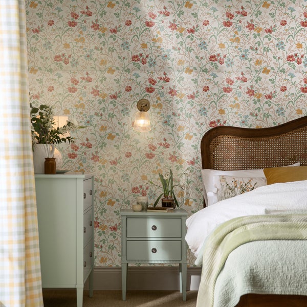 Schlafzimmer mit blumiger Vliestapete, Holz-Bett mit Rattankopfteil, Nachttisch und Wandleuchte in einem hellen, wohnlichen Einrichtungsstil.