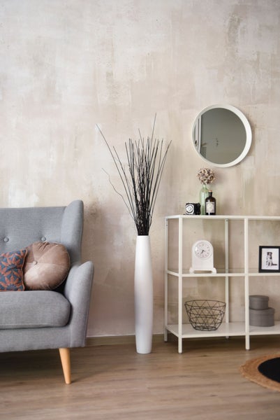 Modernes Wohnzimmer mit grauem Sessel, hoher weißer Bodenvase mit Zweigen, weißem Metallregal und rundem Spiegel vor strukturierter Wand.
