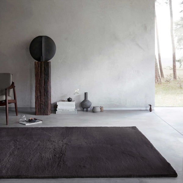 Moderner Wohnbereich mit einem dunkelbraunen Plüschteppich auf Betonboden, einem Holzstuhl und minimalistischer Dekoration vor einer grauen Wand.