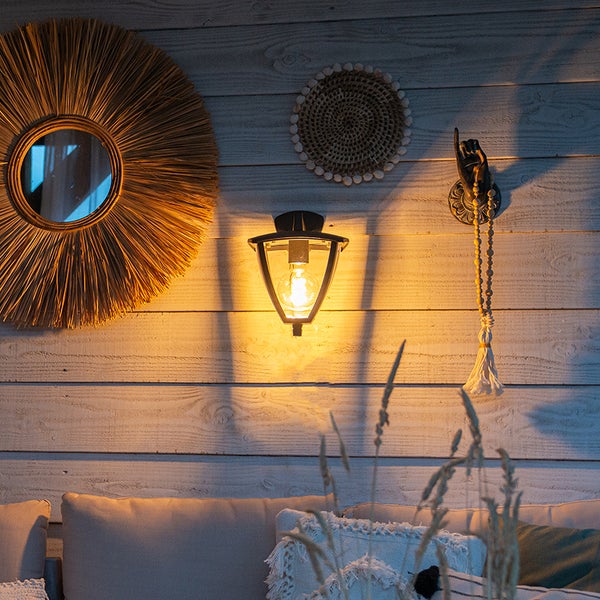 Beleuchtete schwarze Außenwandleuchte an Holzwand neben Sonnenspiegel aus Naturmaterialien und gemütlichen Kissen im Außenbereich.