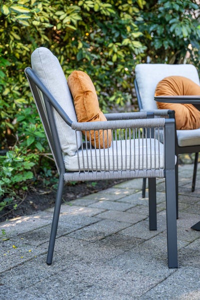 Moderner Gartenstuhl aus dunklem Metall mit hellgrauen Polstern, Seilbespannung an den Seiten und einem orangefarbenen Dekokissen auf einer Terrasse.