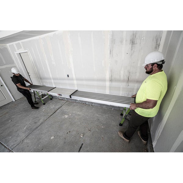 Zwei Handwerker in Schutzkleidung montieren ein langes, ausziehbares Aluminium Arbeitsgerüst auf zwei Böcken in einem unfertigen Innenraum.