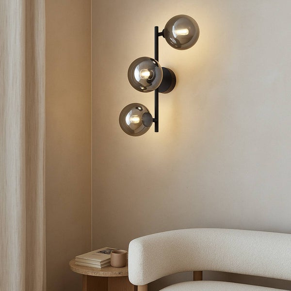 Wandleuchte mit drei Glaskugeln und schwarzem Gestell neben einem Tisch mit Buch und Sessel.