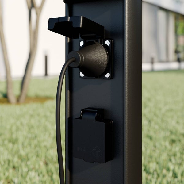 Schwarze Energiesäule aus Metall im hellen Garten mit zwei Steckdosen und Klappdeckeln, eine Steckdose belegt, spritzwassergeschützt.