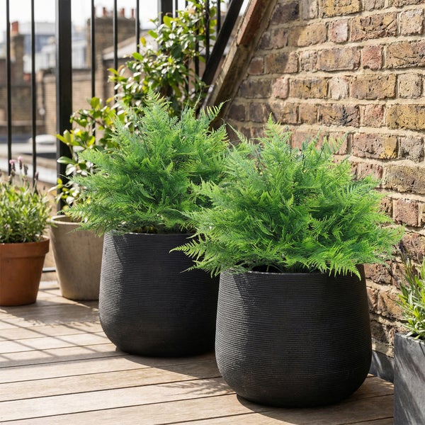 Zwei dunkle, strukturierte Pflanzgefäße mit grünen Farnen auf einer Holzterrasse vor einer Ziegelwand.