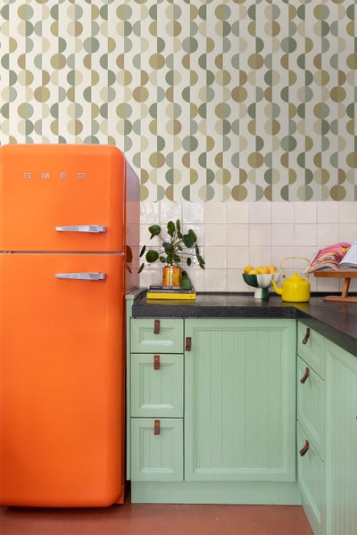 Küche mit geometrischer Retro-Tapete, einem orangefarbenen Smeg-Kühlschrank, minzgrünen Schränken und einer dunklen Arbeitsplatte.