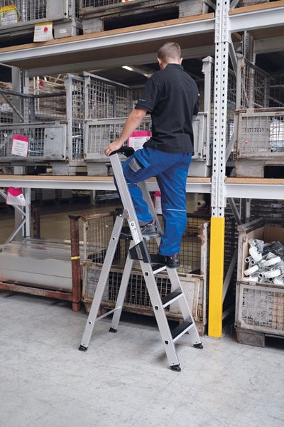 Ein Mitarbeiter steht auf einer stabilen Aluminium Sicherheitsstehleiter in einem Warenlager mit Regalen und Gitterboxen.