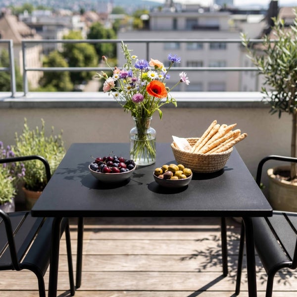Schwarzes Balkonmöbel-Set aus Metall mit quadratischem Tisch und zwei Stühlen, dekoriert mit Blumenvase und Snacks auf einer Holzterrasse.